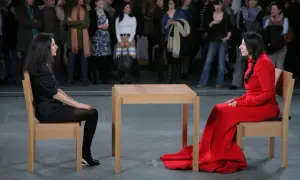Dwie kobiety siedzą naprzeciwko siebie przy drewnianym stole, w ramach performance'u Marina Abramović.