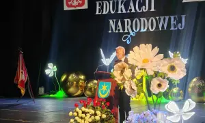 Uroczystość Dzień Edukacji Narodowej. Mężczyzna przemawia, a przed nim bukiety kwiatów. Czy na wernisaż przynosi się kwiaty? Tak, jak widać na scenie!