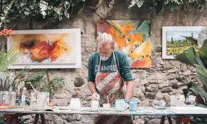 Artysta malarz w fartuchu przygotowuje farby. Obok wiszą jego abstrakcyjne obrazy, pokazując, jak rozlicza się artysta malarz.