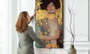 Kobieta wiesza obraz "Judyta z głową Holofernesa" Klimta. Złote zdobienia i mroczna atmosfera dzieła przyciągają wzrok.