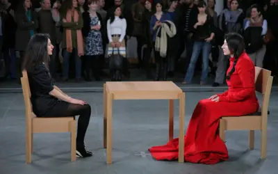 Dwie kobiety siedzą naprzeciwko siebie przy drewnianym stole, w ramach performance'u Marina Abramović.