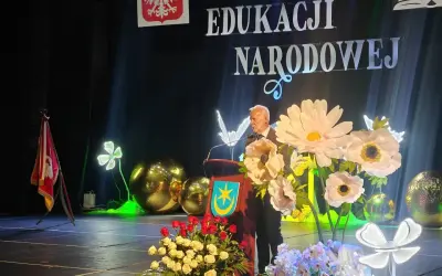 Uroczystość Dzień Edukacji Narodowej. Mężczyzna przemawia, a przed nim bukiety kwiatów. Czy na wernisaż przynosi się kwiaty? Tak, jak widać na scenie!