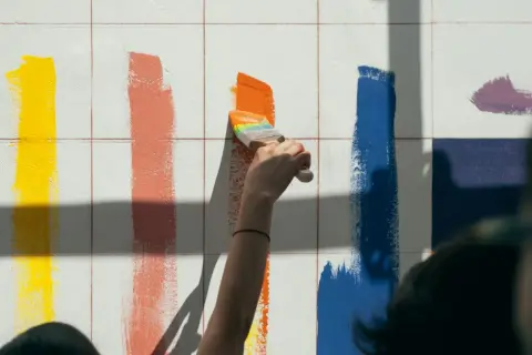 Ręka z pędzlem maluje pomarańczowy pas na białej ścianie. Obok żółte, koralowe i niebieskie paski. Technika malarska w akcji.