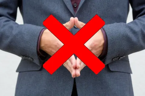 Postać w sztuce, mężczyzna w garniturze, z dłońmi złożonymi na piersi, na które nałożono duży czerwony znak "X".