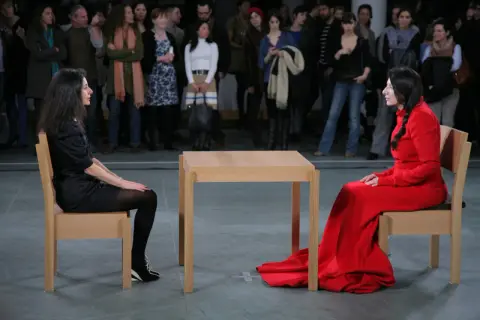 Dwie kobiety siedzą naprzeciwko siebie przy drewnianym stole, w ramach performance'u Marina Abramović.