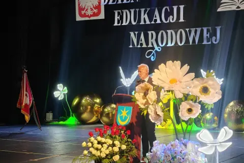 Uroczystość Dzień Edukacji Narodowej. Mężczyzna przemawia, a przed nim bukiety kwiatów. Czy na wernisaż przynosi się kwiaty? Tak, jak widać na scenie!