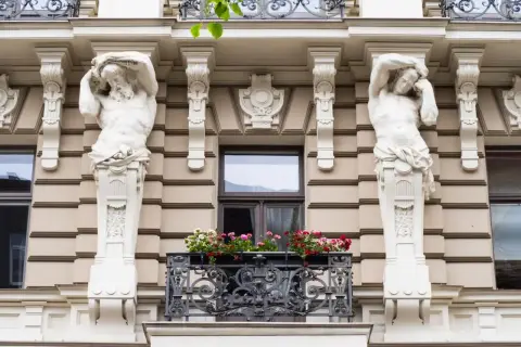 Dwie rzeźby atlantesów podtrzymują balkon w stylu secesyjnym. Kwiaty dodają koloru tej architekturze.