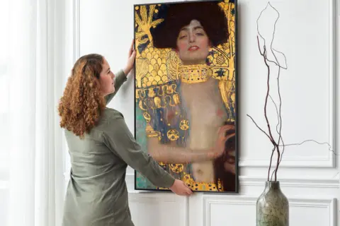 Kobieta wiesza obraz "Judyta z głową Holofernesa" Klimta. Złote zdobienia i mroczna atmosfera dzieła przyciągają wzrok.
