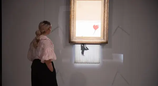 Kobieta ogląda słynne dzieło Banksy'ego, które uległo samozniszczeniu. Obraz z czerwonym balonem w kształcie serca jest częściowo zniszczony.