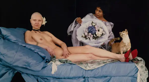 Katarzyna Kozyra w "Łaźni" - naga kobieta z kwiatem we włosach leży na niebieskiej pościeli, obok kot i bukiet.