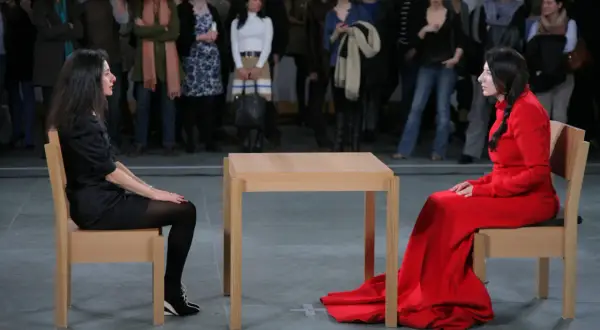 Dwie kobiety siedzą naprzeciwko siebie przy drewnianym stole, w ramach performance'u Marina Abramović.