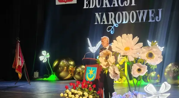 Uroczystość Dzień Edukacji Narodowej. Mężczyzna przemawia, a przed nim bukiety kwiatów. Czy na wernisaż przynosi się kwiaty? Tak, jak widać na scenie!