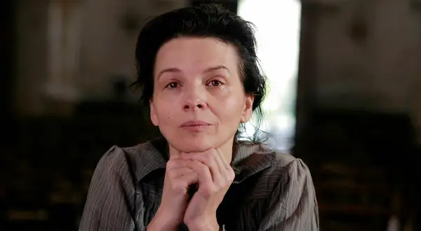 Portret kobiety w wieku dojrzałym, przypominającej Camille Claudel, z dłońmi podpartymi pod brodą, w stonowanym wnętrzu.