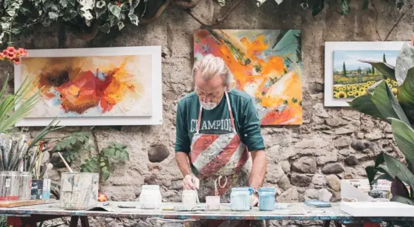 Artysta malarz w fartuchu przygotowuje farby. Obok wiszą jego abstrakcyjne obrazy, pokazując, jak rozlicza się artysta malarz.