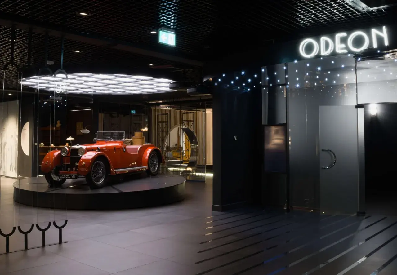 Pomarańczowy kabriolet z lat 20. XX wieku, symbol sztuki dwudziestolecia międzywojennego, prezentowany w nowoczesnym wnętrzu z neonowym napisem "ODEON".