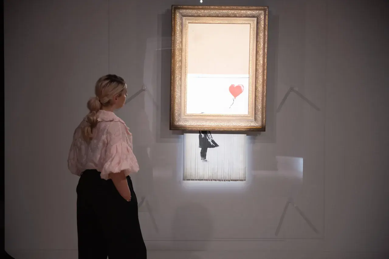 Kobieta ogląda słynne dzieło Banksy'ego, które uległo samozniszczeniu. Obraz z czerwonym balonem w kształcie serca jest częściowo zniszczony.