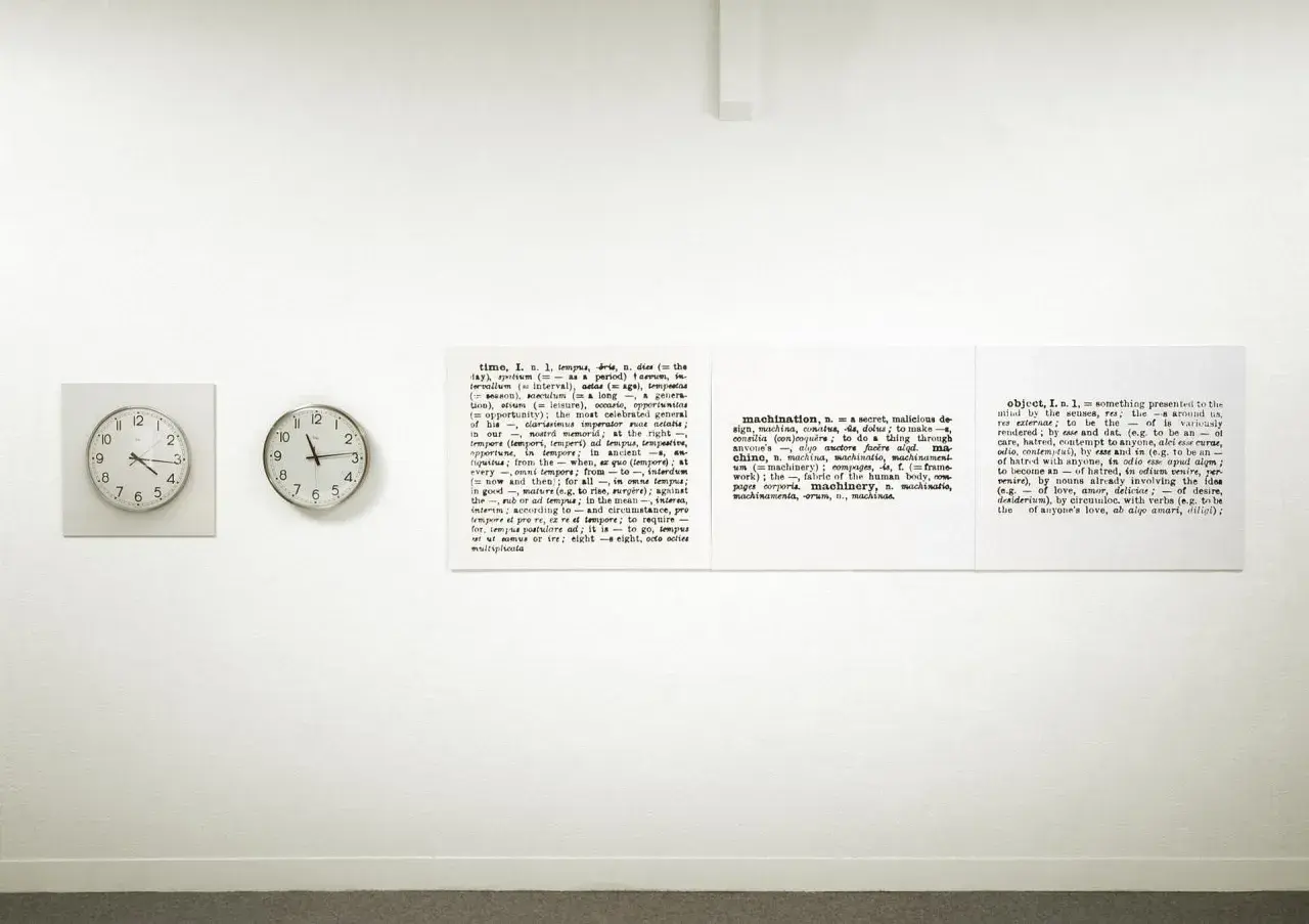 Sztuka konceptualna: dwa zegary i słownikowe definicje słów "time", "machination" i "object" na białej ścianie.