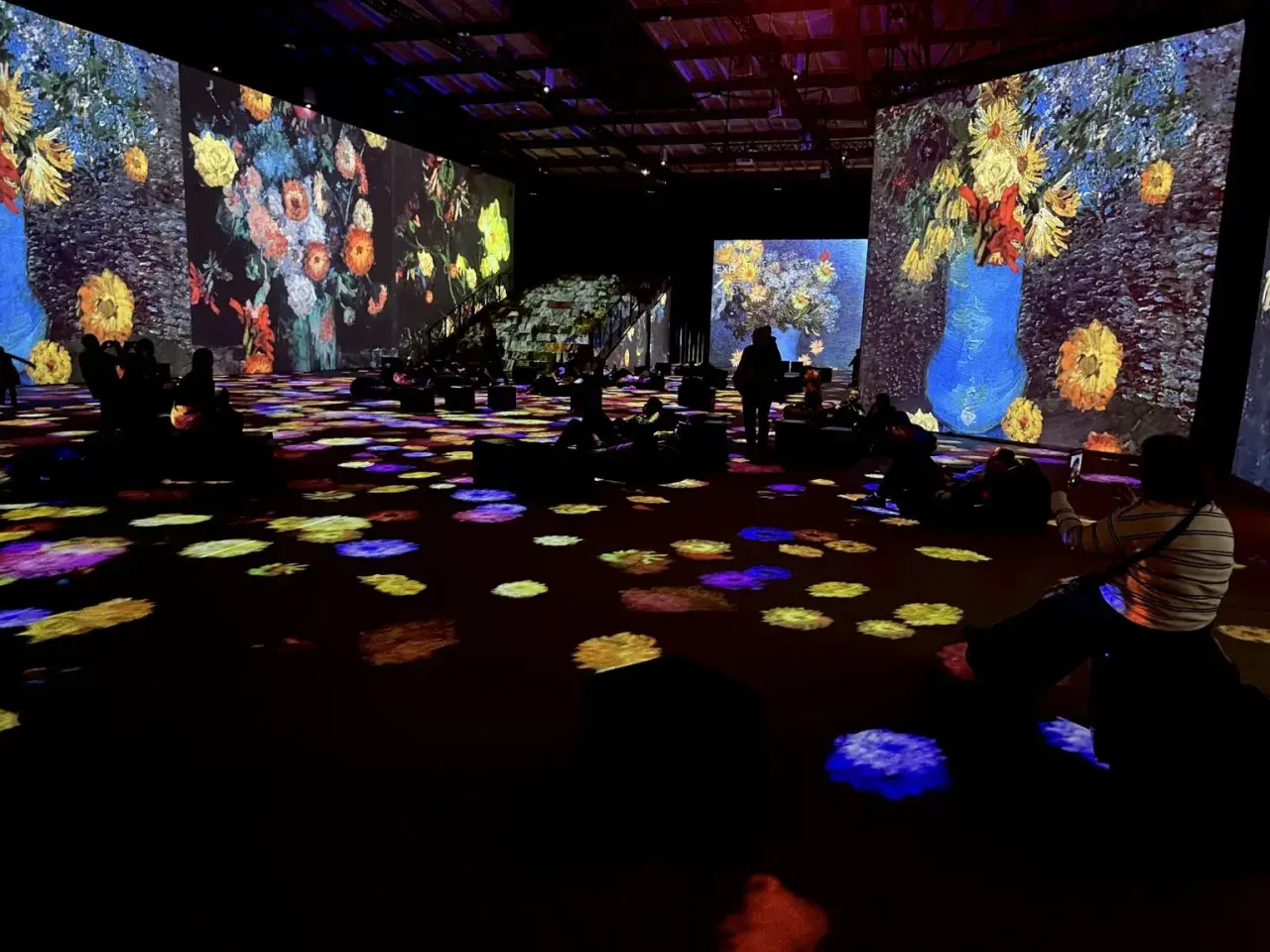 Wystawa Van Gogh: widzowie zanurzeni w projekcjach słoneczników i innych dzieł, siedzą na podłodze oświetlonej kolorowymi plamami.