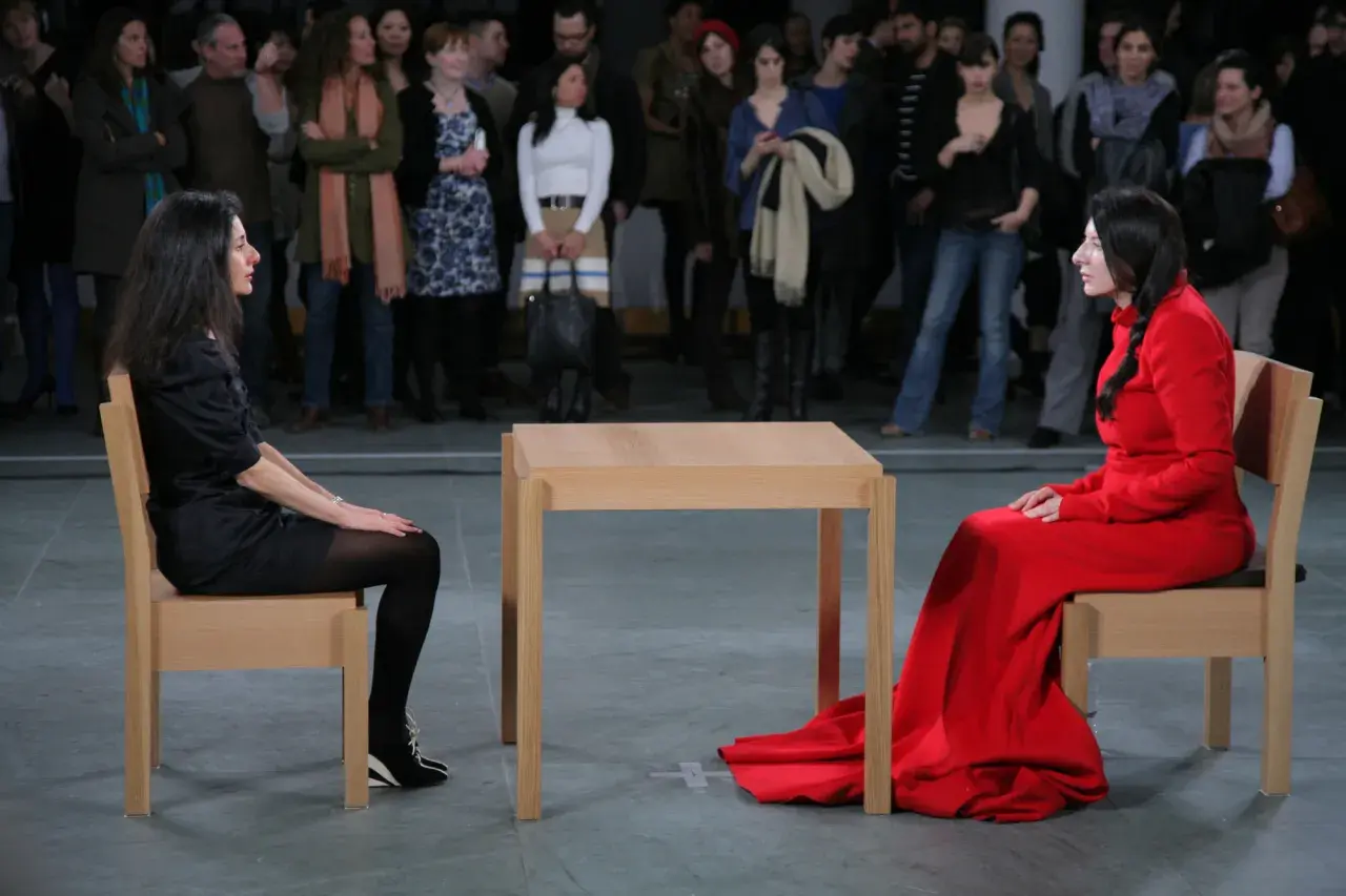 Dwie kobiety siedzą naprzeciwko siebie przy drewnianym stole, w ramach performance'u Marina Abramović.