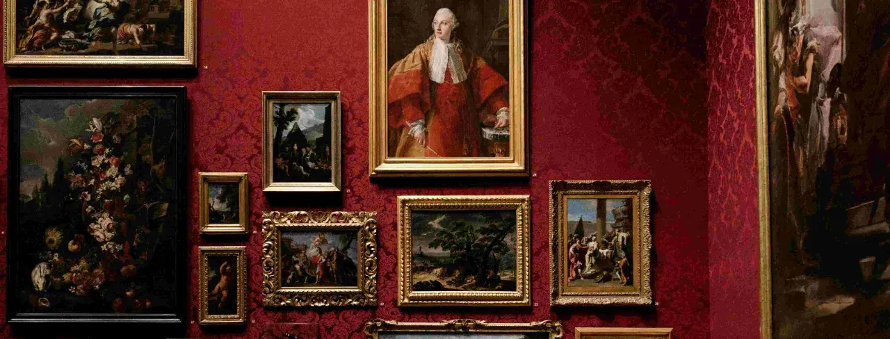 Galeria sztuki z obrazami w złotych ramach na czerwonej, wzorzystej ścianie. Widoczny portret mężczyzny w stroju z epoki, martwa natura z kwiatami i sceny rodzajowe. Różnorodność stylów ukazuje bogactwo nurtów w sztuce.