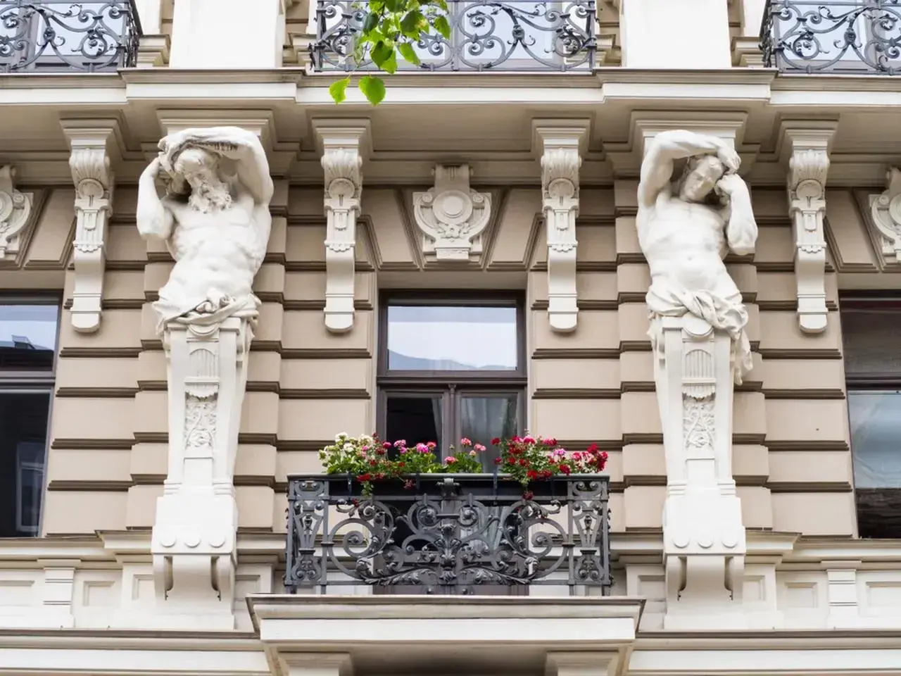 Dwie rzeźby atlantesów podtrzymują balkon w stylu secesyjnym. Kwiaty dodają koloru tej architekturze.