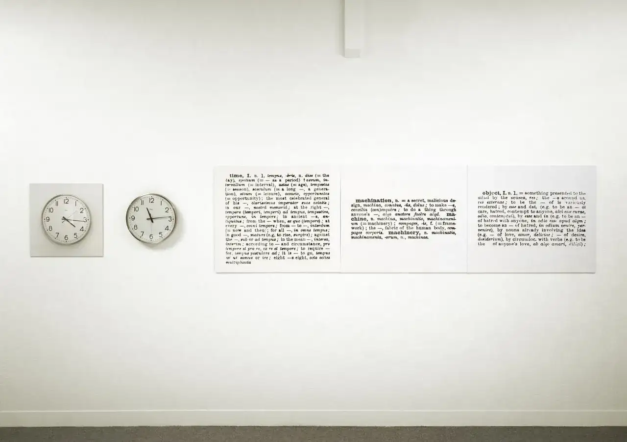 Sztuka konceptualna: dwa zegary i słownikowe definicje słów "time", "machination" i "object" na białej ścianie.