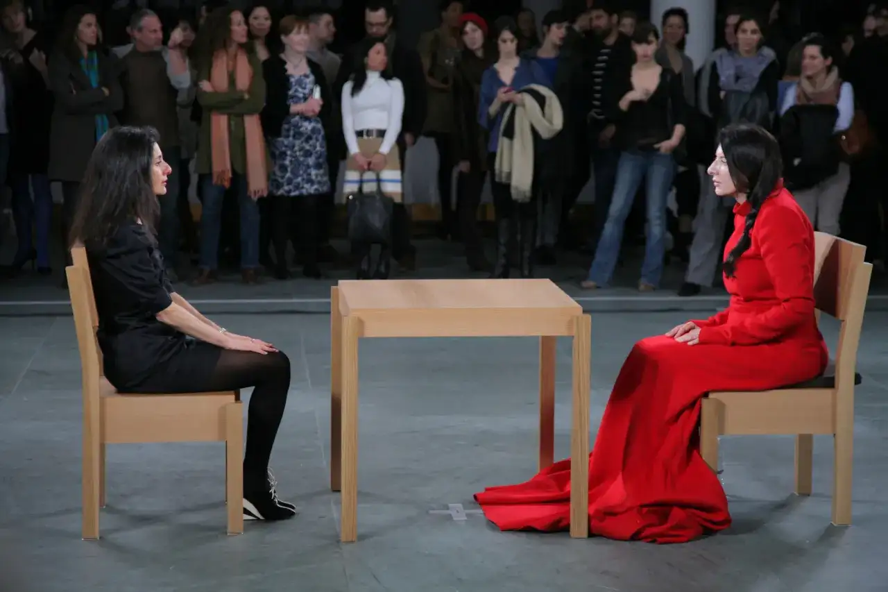 Dwie kobiety siedzą naprzeciwko siebie przy drewnianym stole, w ramach performance'u Marina Abramović.