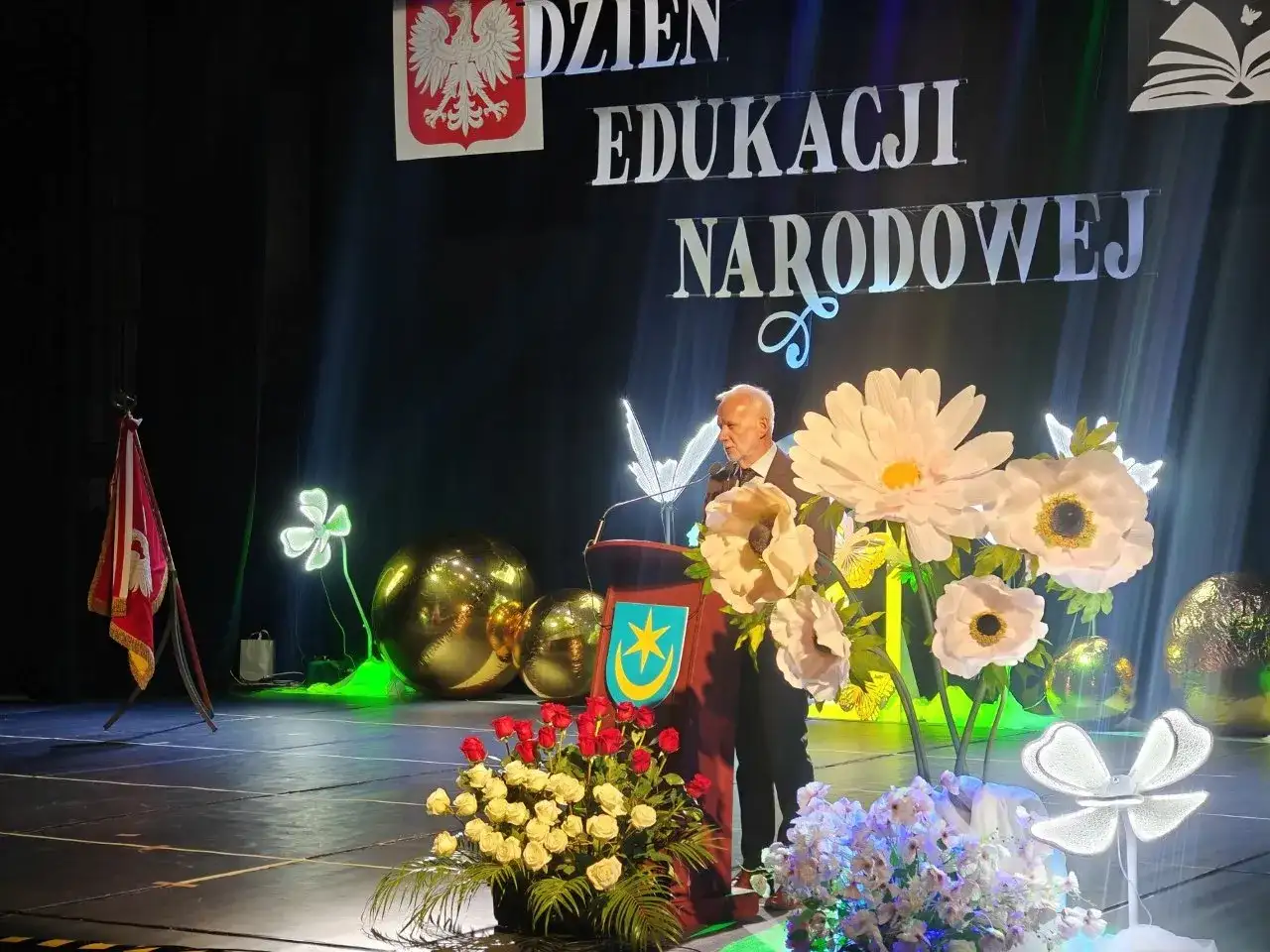 Uroczystość Dzień Edukacji Narodowej. Mężczyzna przemawia, a przed nim bukiety kwiatów. Czy na wernisaż przynosi się kwiaty? Tak, jak widać na scenie!