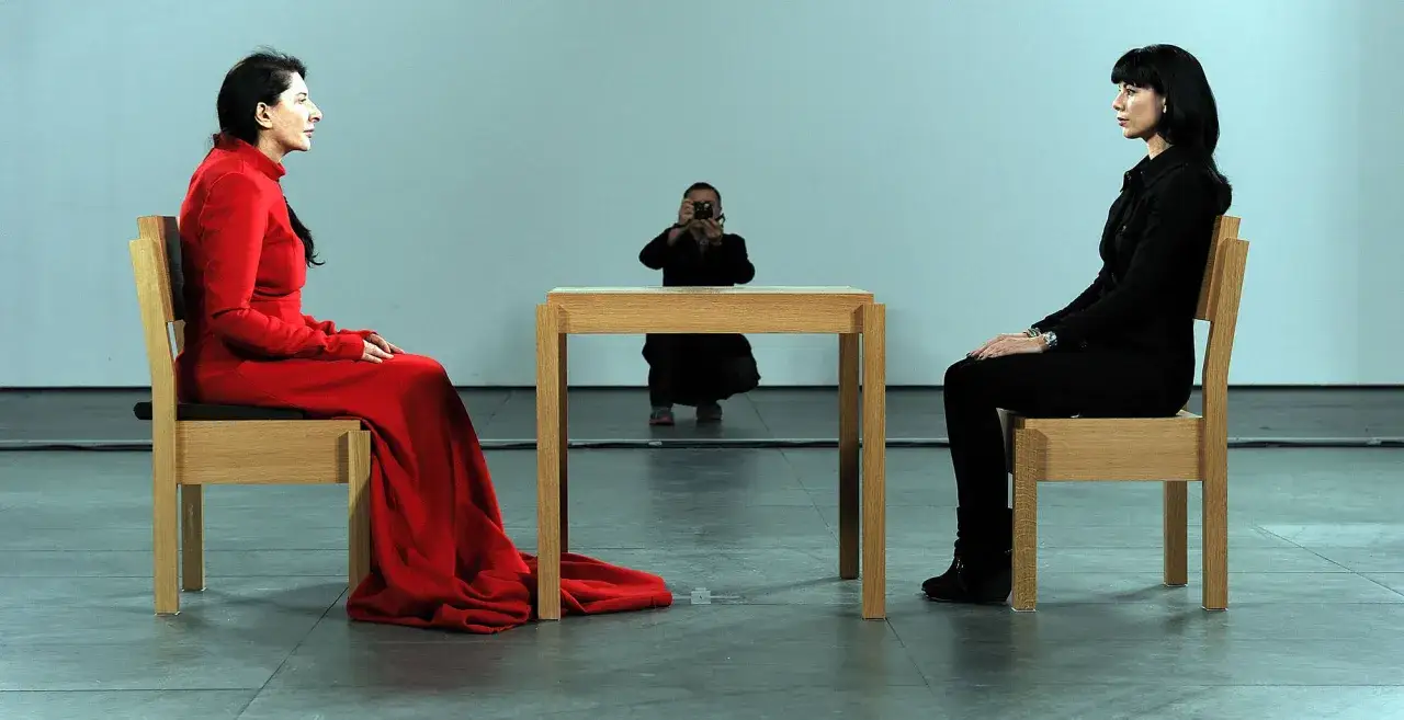 Dwie kobiety siedzą naprzeciwko siebie. Jedna w czerwieni, druga w czerni. W tle osoba robiąca zdjęcie. Ikoniczna marina abramovic performance.