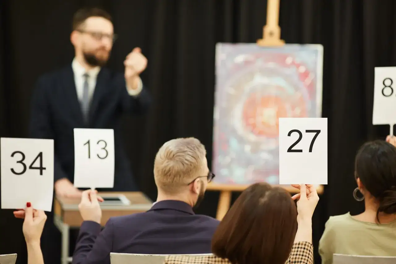 Licytacja dzieła sztuki. Aukcjoner zachęca do podbijania cen, a widzowie podnoszą tabliczki z numerami. Wśród nich jest potencjalny mecenas sztuki.