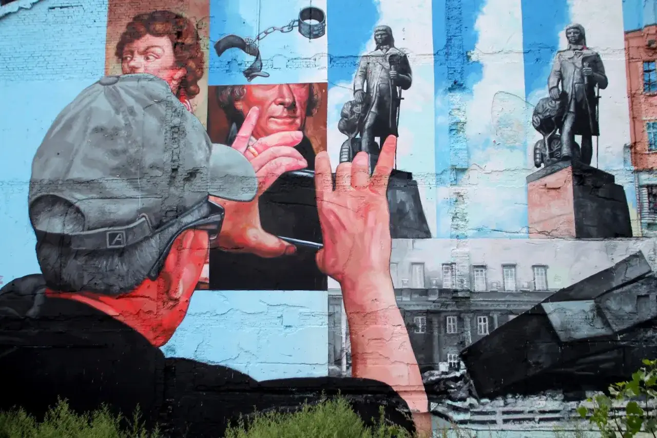 Co to jest mural? Dzieło sztuki na ścianie przedstawia człowieka w czapce i okulary, który patrzy na portrety i pomniki.