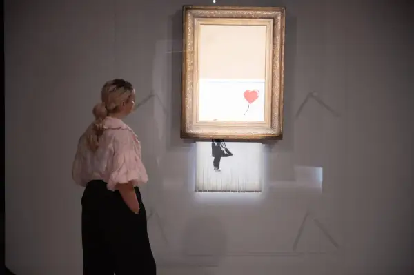 Kobieta ogląda słynne dzieło Banksy'ego, które uległo samozniszczeniu. Obraz z czerwonym balonem w kształcie serca jest częściowo zniszczony.