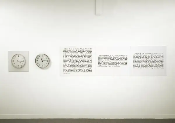 Sztuka konceptualna: dwa zegary i słownikowe definicje słów "time", "machination" i "object" na białej ścianie.