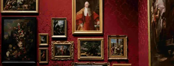 Galeria sztuki z obrazami w złotych ramach na czerwonej, wzorzystej ścianie. Widoczny portret mężczyzny w stroju z epoki, martwa natura z kwiatami i sceny rodzajowe. Różnorodność stylów ukazuje bogactwo nurtów w sztuce.