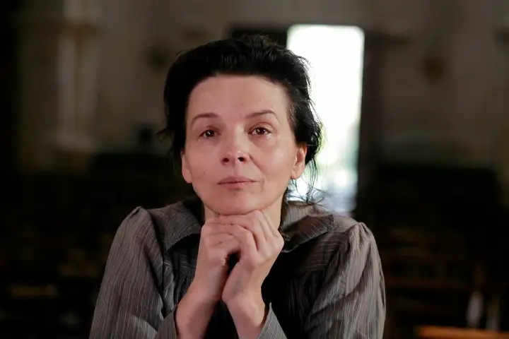 Portret kobiety w wieku dojrzałym, przypominającej Camille Claudel, z dłońmi podpartymi pod brodą, w stonowanym wnętrzu.